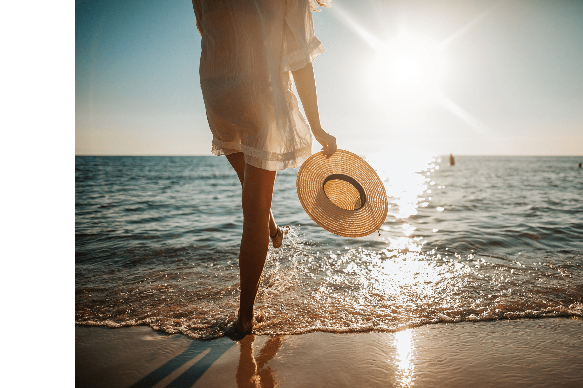 Close-up of young woman in white sun dress and with hat in hand walking alone on sandy beach at summer sunset, splashing water in sea shallow
