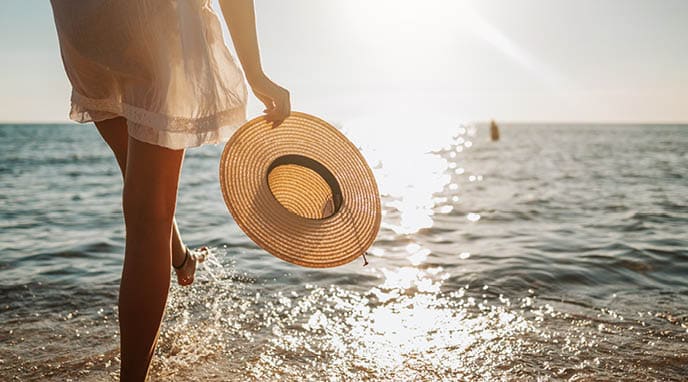 Close-up of young woman in white sun dress and with hat in hand walking alone on sandy beach at summer sunset, splashing water in sea shallow