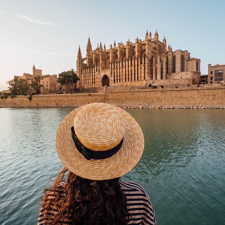 Rear view of a woman with a straw hat while she's admiring the Cathedral de Santa Mar a de Palma de Mallorca at sunset. The Cathedral reflects in the water.