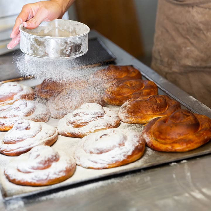 Baker hands dusting freshly baked ensaimadas with powdered sugar in bakery. Baking process of traditional Spanish pastries