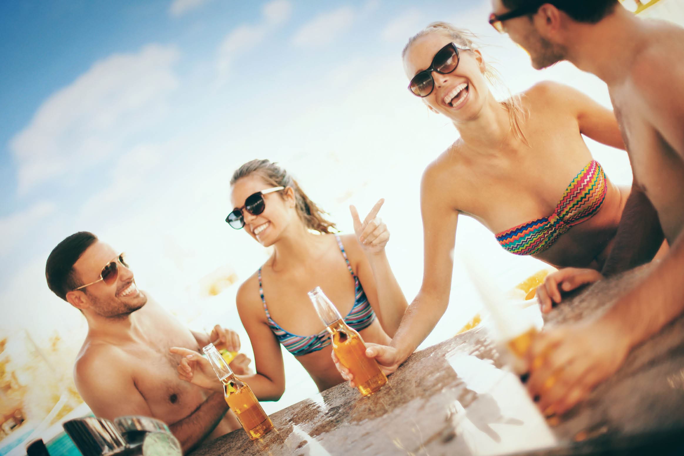 Group of mid 20's couples having fun at a beach bar. They are dancing and drinking beers. There are two guys and two girls, blurry swimming pool behind them. Each person wearing sunglasses.