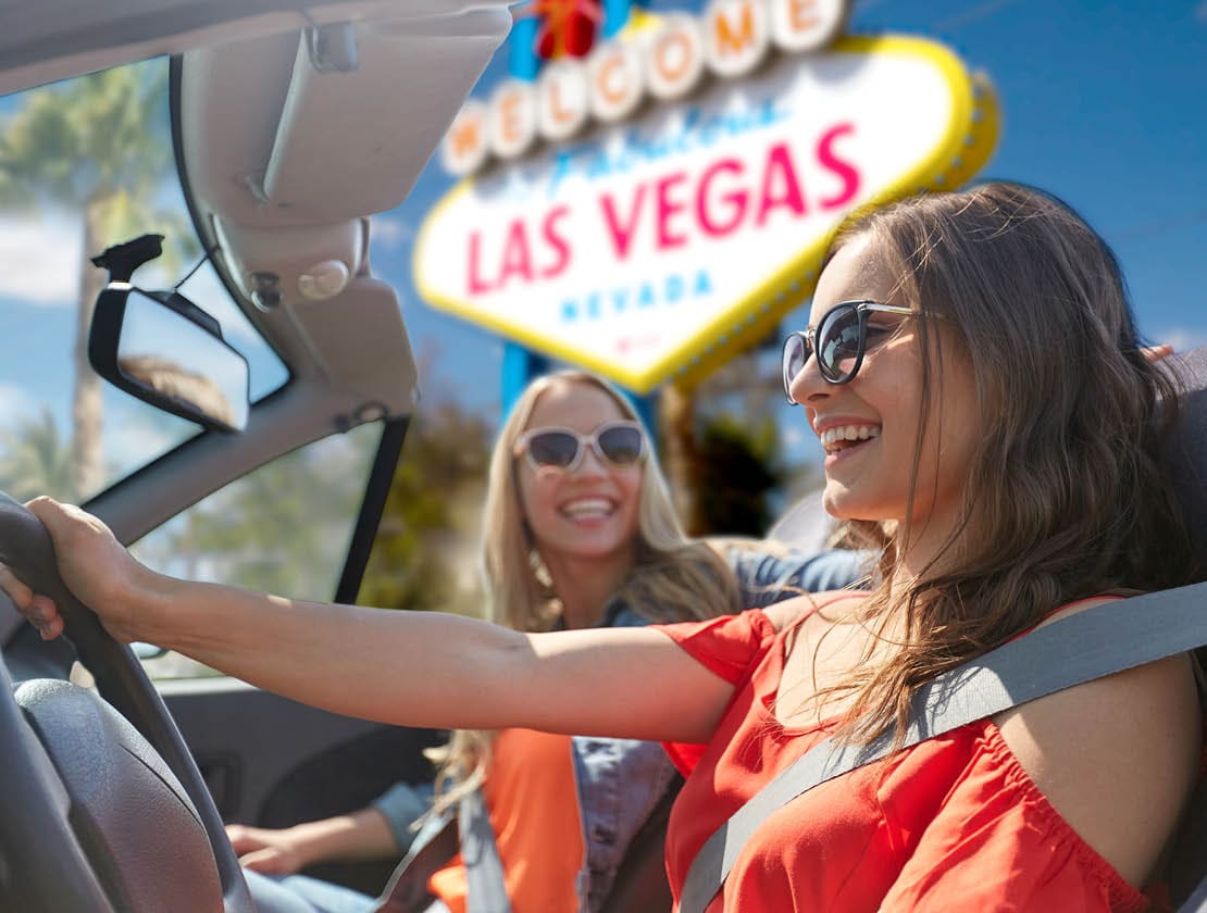 summer holidays, road trip and travel concept - happy young women driving in convertible car and laughing over welcome to fabulous las vegas sign background